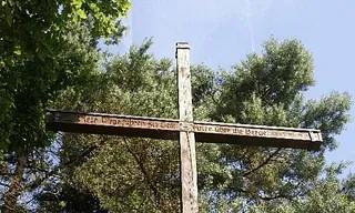 Großes Holzkreuz mit eingraviertem Text vor einem Baum.