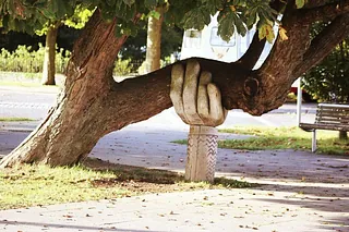 Skulptur einer Hand, die eine geflochtene Säule hält, unter einem Baum.